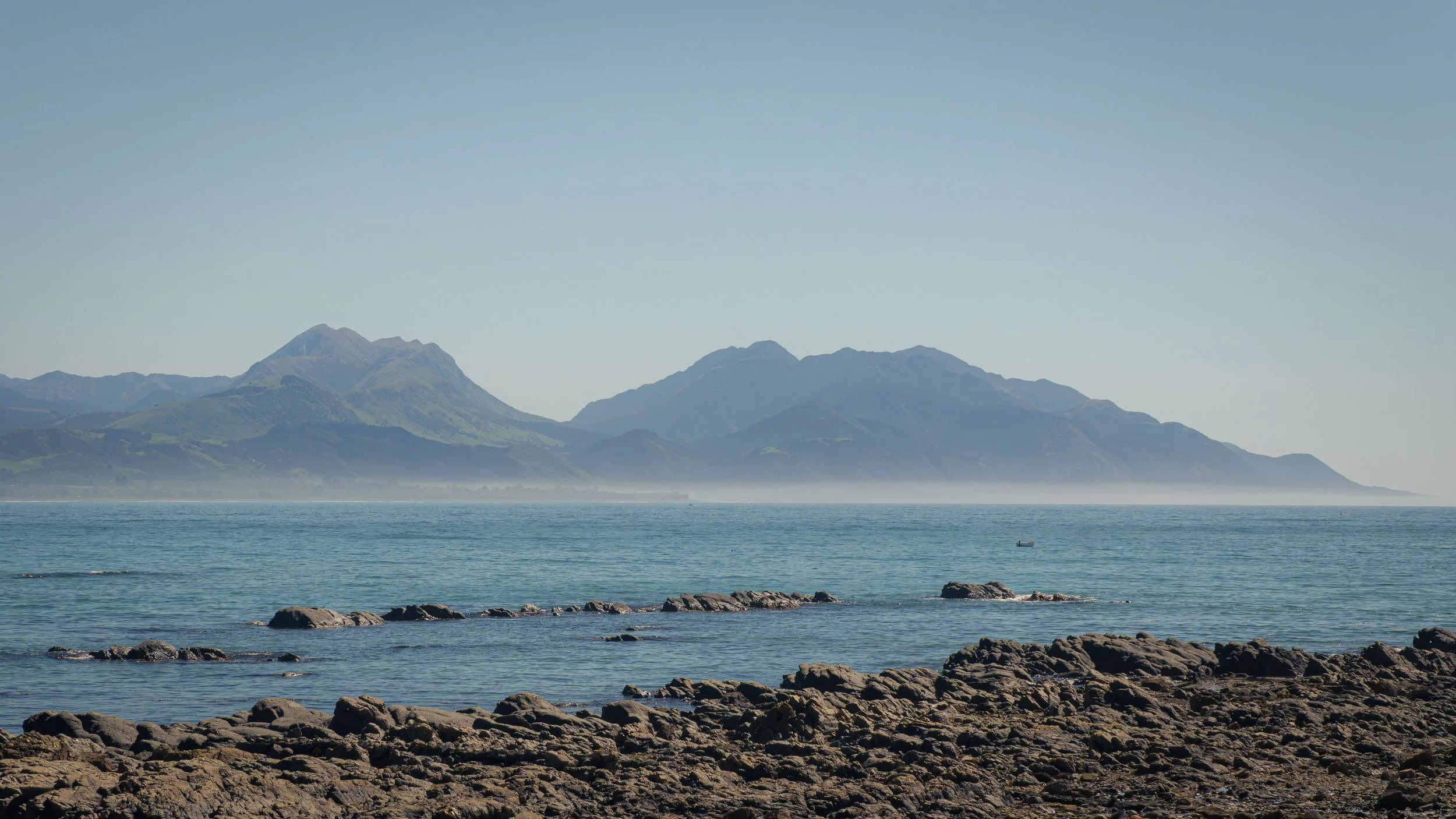 View across the rocky Kaikōura coastline towards the Kaikōura Ranges, with calm ocean waters and distant mountain peaks under clear skies.