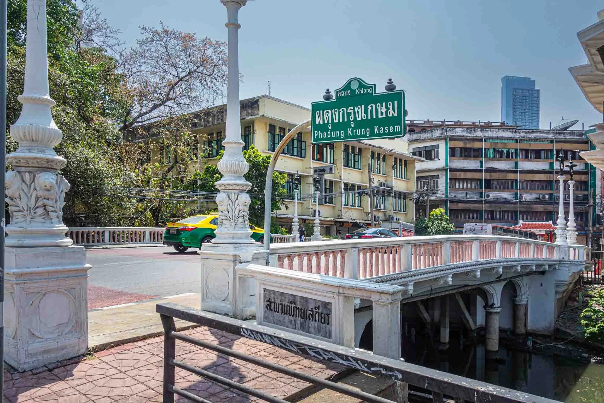 Street scene showing a green and yellow taxi on a road with buildings in the background. There are street signs in Thai and English indicating the location as Phadung Krung Kasem in Bangkok, Thailand. The scene includes decorative white railings and 