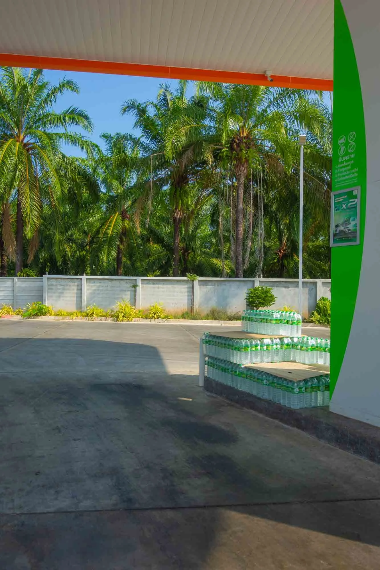 A gas station canopy with a mountain of bottled water stacked on a small platform outside, with lush green palm trees and a concrete fence in the background.