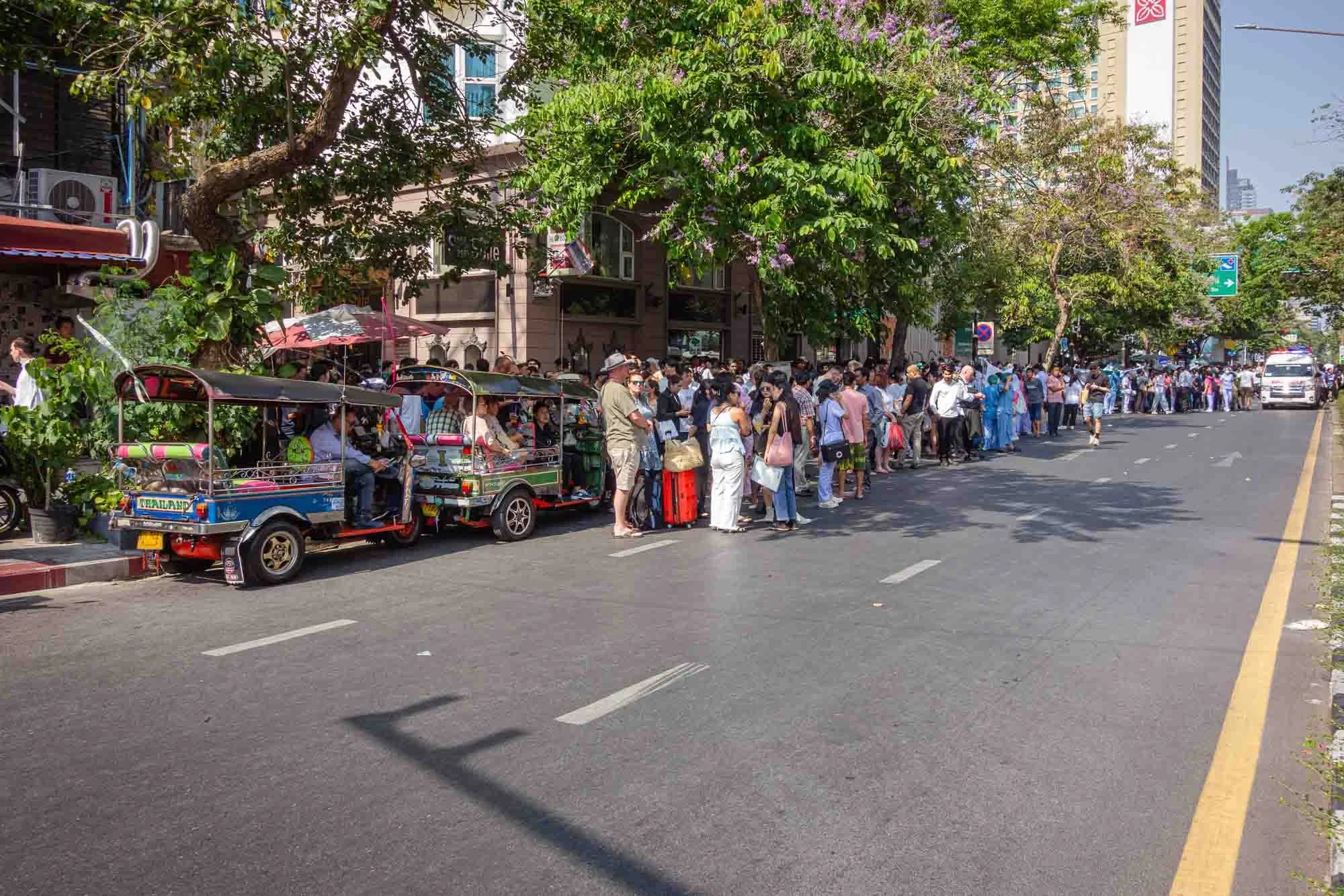 Long line of people waiting outside a building, with trees and city buildings in the background, and a small vehicle parked on the street.