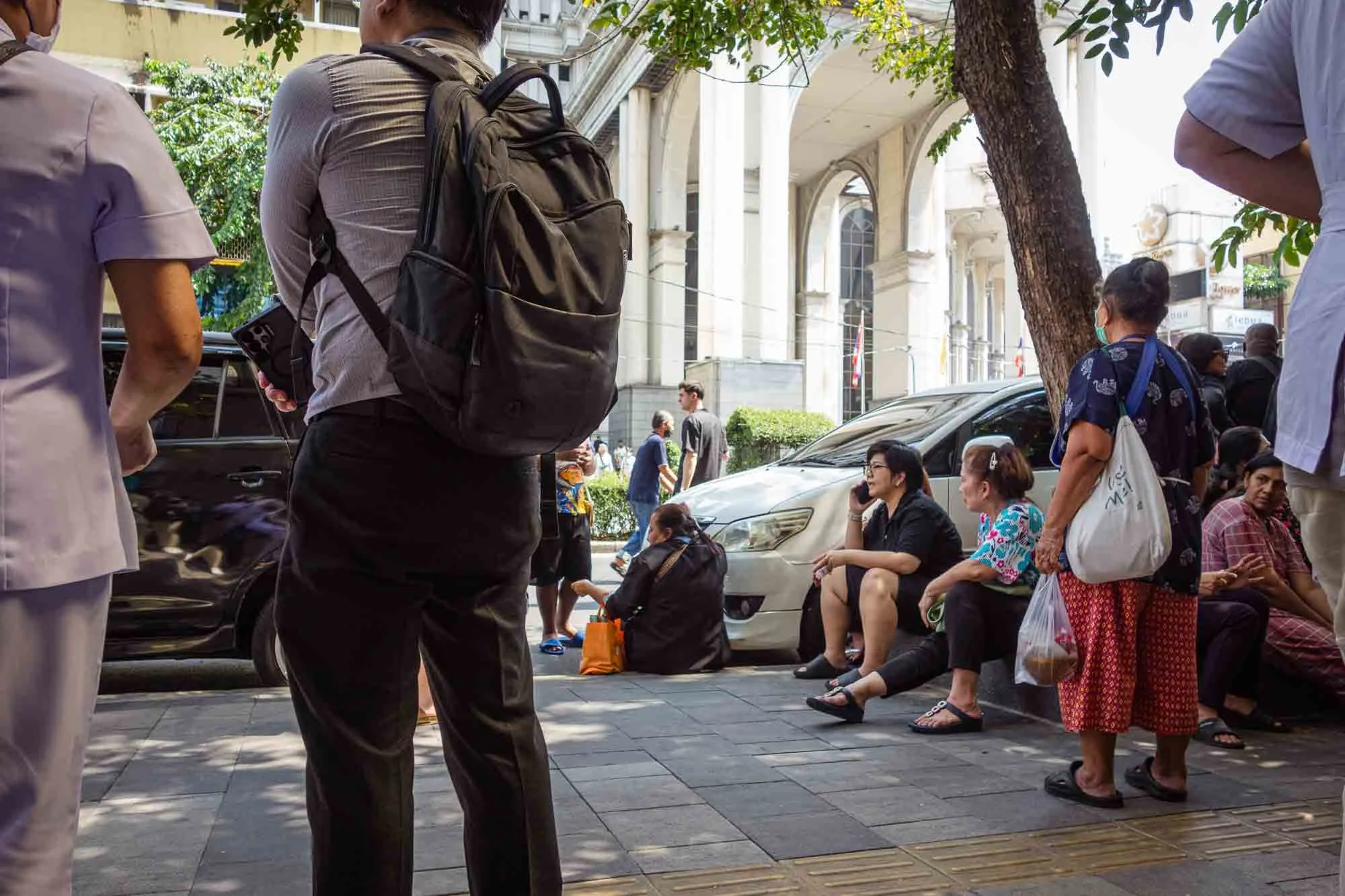 People waiting and sitting on the sidewalk near a tree and parked cars, with a historic building in the background.