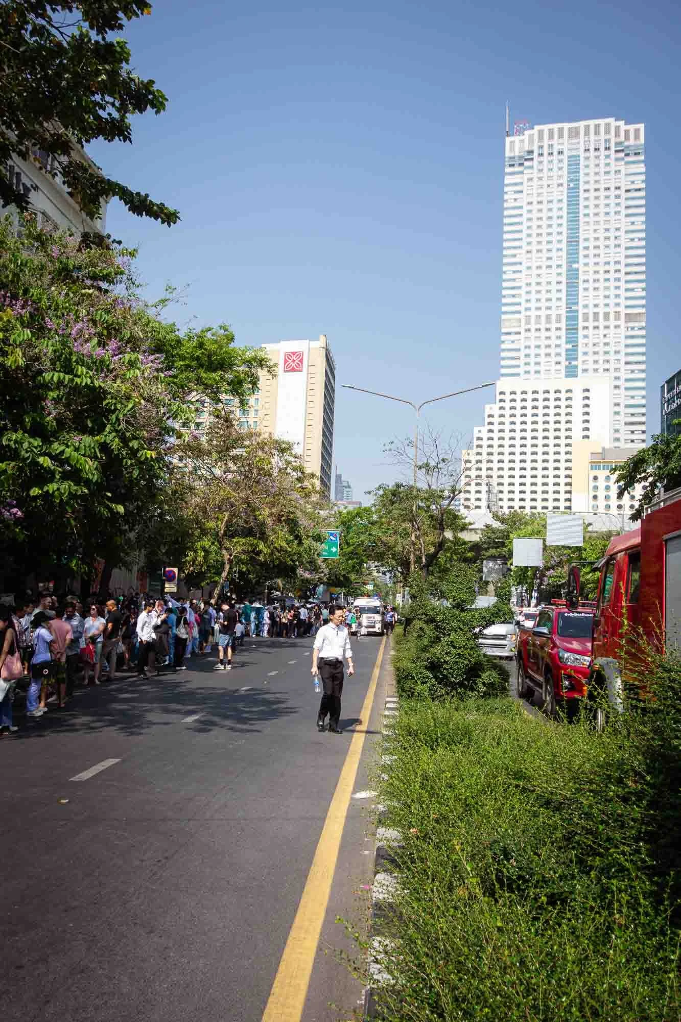 City street with a crowd of people waiting to cross, tall buildings in the background, and vehicles on the road, sunny day.