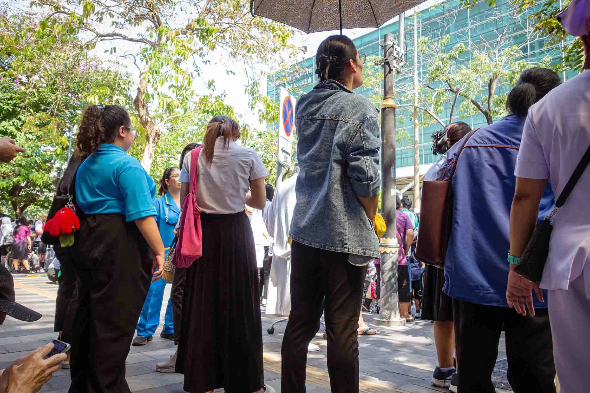 Crowd of people standing on a sidewalk under trees in front of a modern building with glass windows; some are wearing uniforms and face masks.
