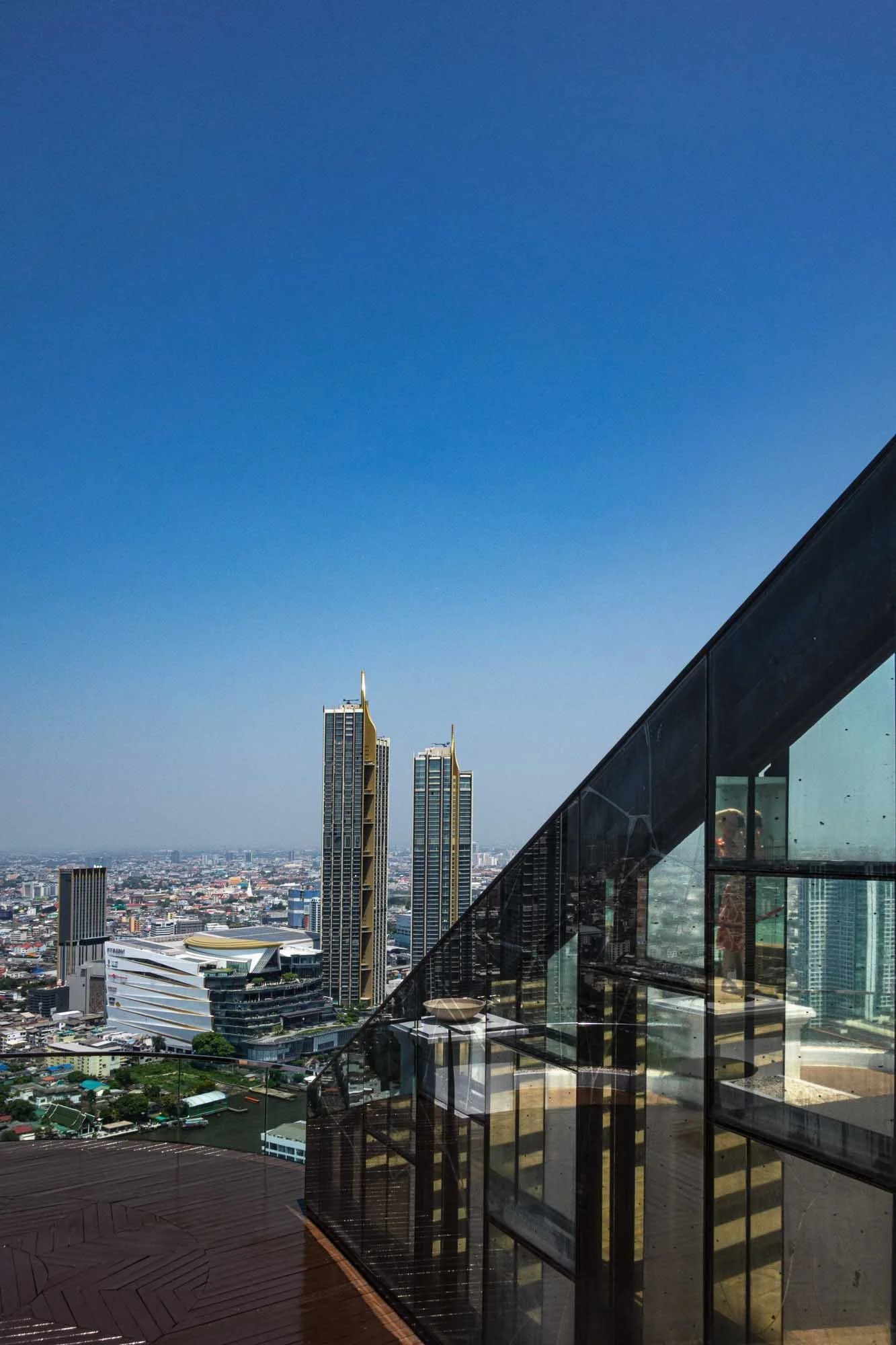 City skyline with two tall modern skyscrapers with gold accents, viewed from a balcony with glass railings and wood decking, under a clear blue sky.