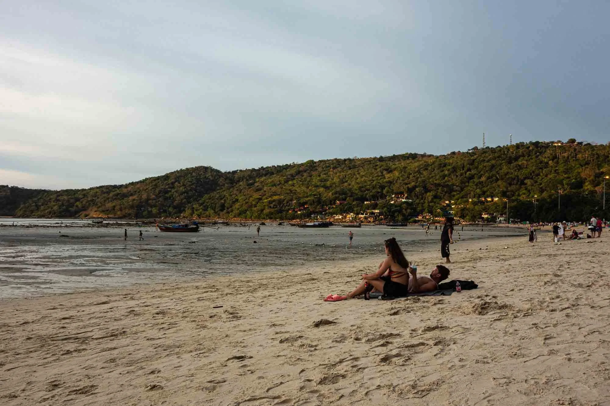 People relaxing on a sandy beach with green hills and a cloudy sky in the background.