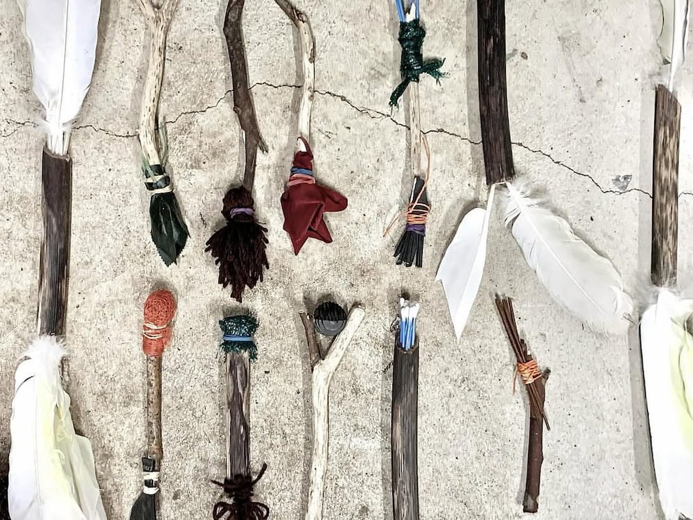 Collection of handmade painting sticks decorated with feathers, yarn, and fabric, arranged on a concrete surface. Ready to be used as paint brushes and mark making