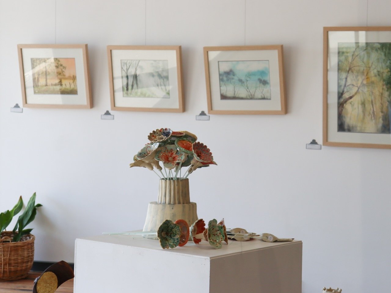 An art gallery with four framed landscape watercolor paintings hung on a white wall, a display of ceramic flower sculptures on a white pedestal, and a potted plant in a wicker basket on the floor.