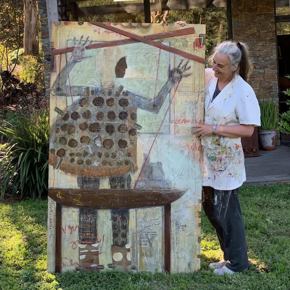 Macedon Ranges Artist Sam Gardiner with one of her mixed media paintings. The painting is propped up against a building with stone siding, and the woman is smiling while holding the painting.