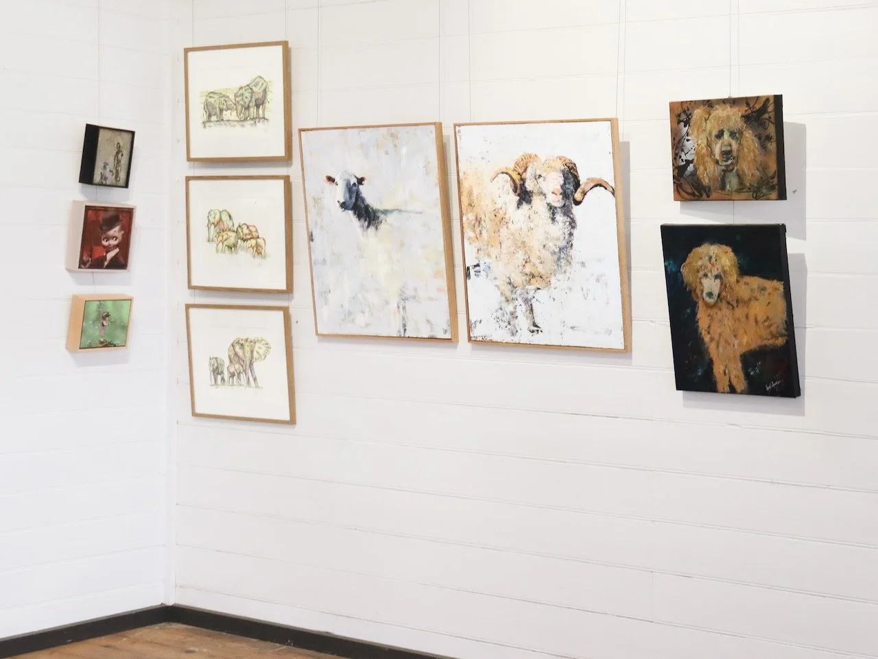 Gallery wall with various animal paintings, including several of sheep, goats, a dog, and a lion, framed and displayed on a white wood-paneled wall.