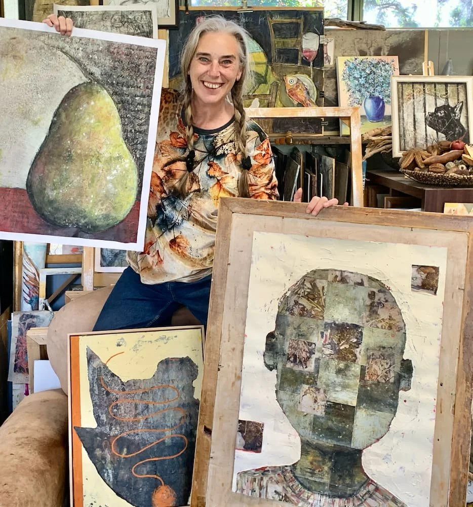 Sam Gardiner, wearing a floral shirt, smiling and holding two framed pieces of artwork. The artwork features a pear and a collage portrait of a person's face. The background shows a room filled with various paintings, paintings, and art supplies.