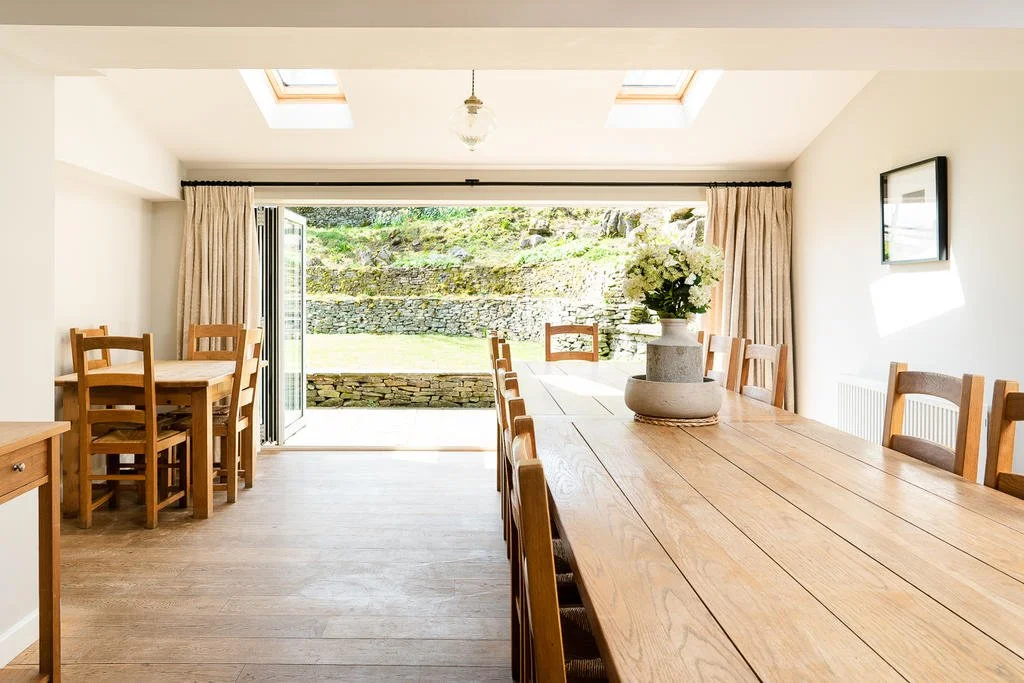 Sunlit dining room with large wooden table, chairs, and a vase of white flowers, opening to a patio with a stone wall and greenery outside.
