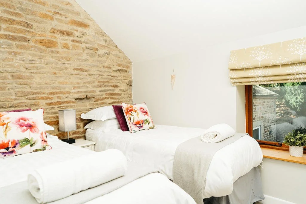 Two beds with white bedding, decorative pillows, a nightstand with a lamp, and a window with a sunlight view in a bedroom with a stone accent wall.