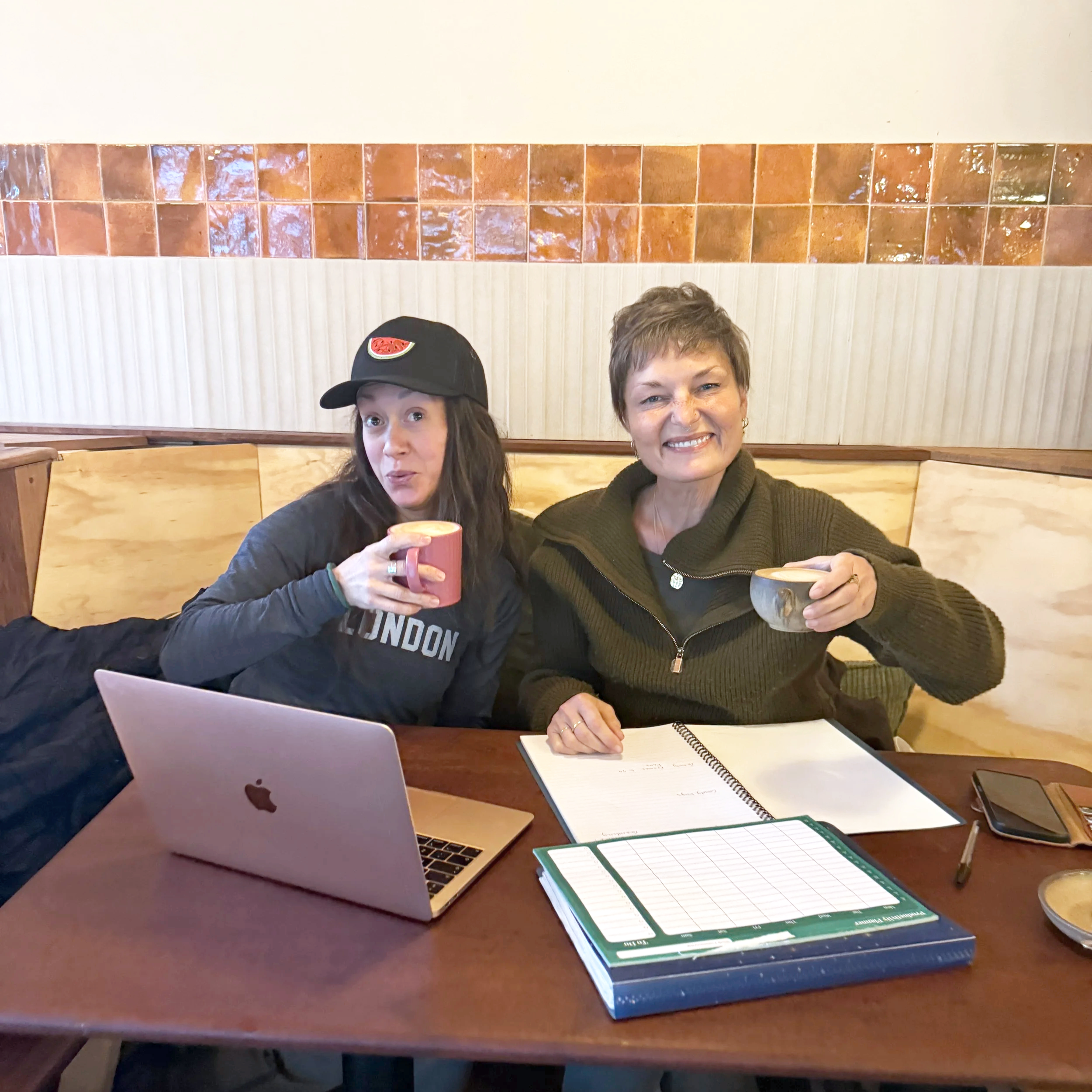 Two women sitting at a table in a casual restaurant, smiling and holding cups of coffee. The woman on the left is wearing a black baseball cap with a watermelon logo, a black shirt that says "LONDON," and is using a laptop. The woman on the right has short hair, is wearing a dark green zip-up sweater, and has a notebook, clipboard, and smartphone in front of her.