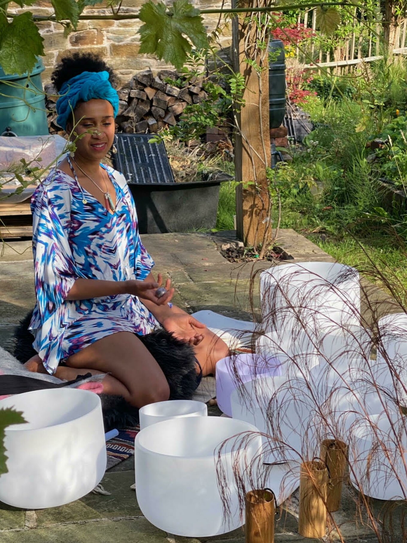 A woman in a colorful dress and blue headwrap is sitting outdoors on a rug, playing a singing bowl with a mallet, surrounded by decorative glass and metal art pieces, in a garden setting with plants and a brick wall in the background.