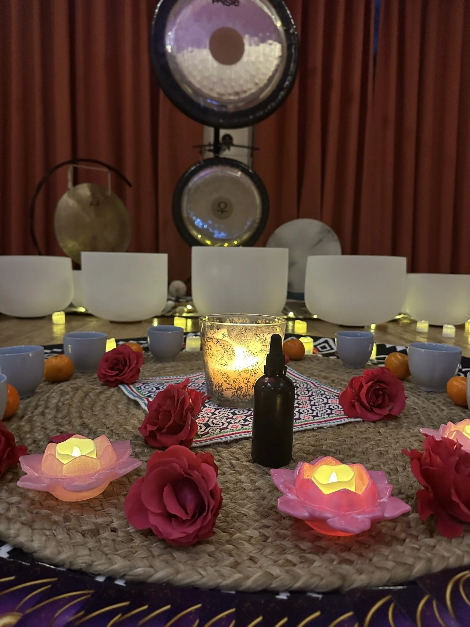 A decorated table with a candle holder, pink roses, small candles, cups, and fruits, set against a backdrop of musical instruments including gongs.