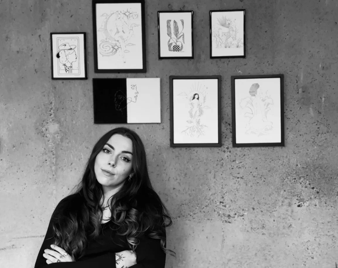 A woman with long hair and a nose piercing standing with arms crossed in front of a concrete wall decorated with eight framed art prints, including drawings of a person, plants, and abstract designs.