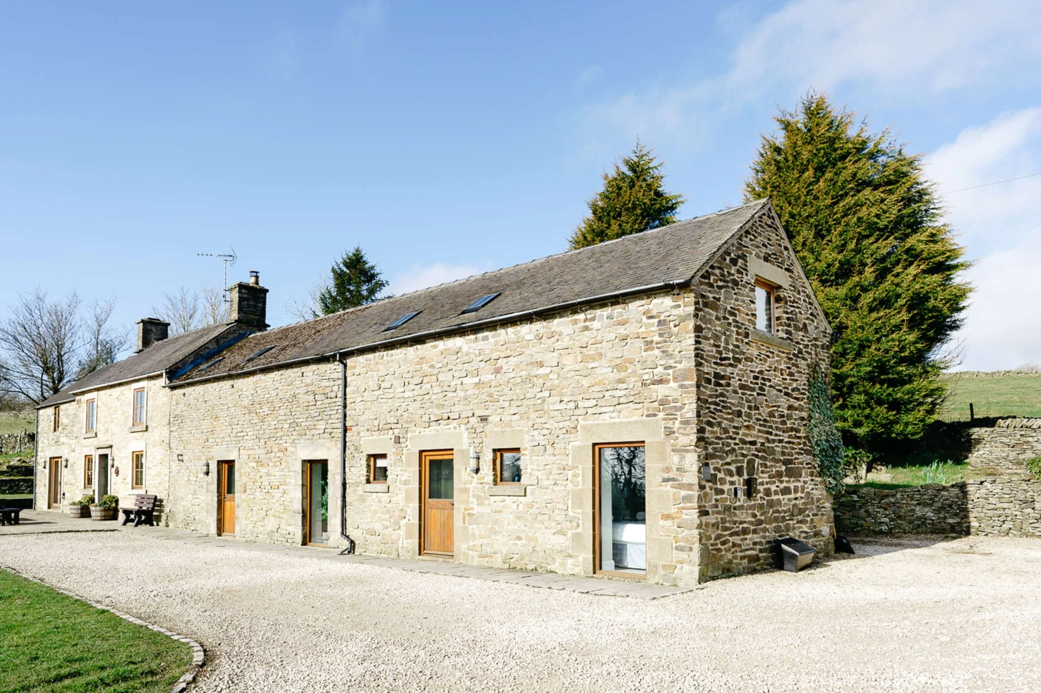 Lapwing Barns, a Stone farmhouse with wooden doors and windows, gravel driveway, surrounded by trees and green grass on a sunny day.