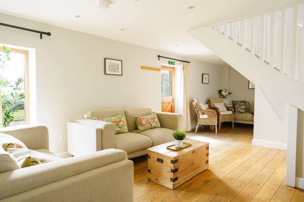 Bright living room with white walls, wooden floor, beige sofas with floral cushions, a wooden coffee table, wicker chairs with cushions, and a staircase with white railing.