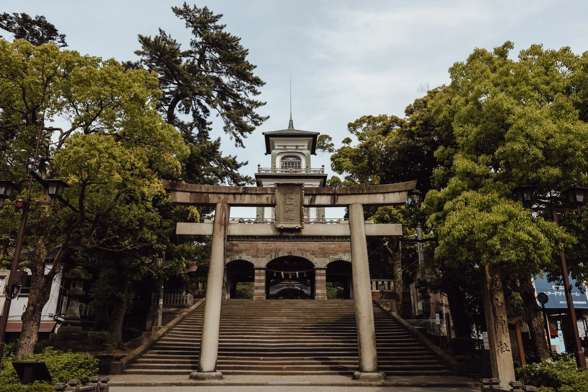 A concrete torii gate in front of the unique dutch and japanese architectural gate at Oyama shrine surrounded by lush large green trees under a blue sky