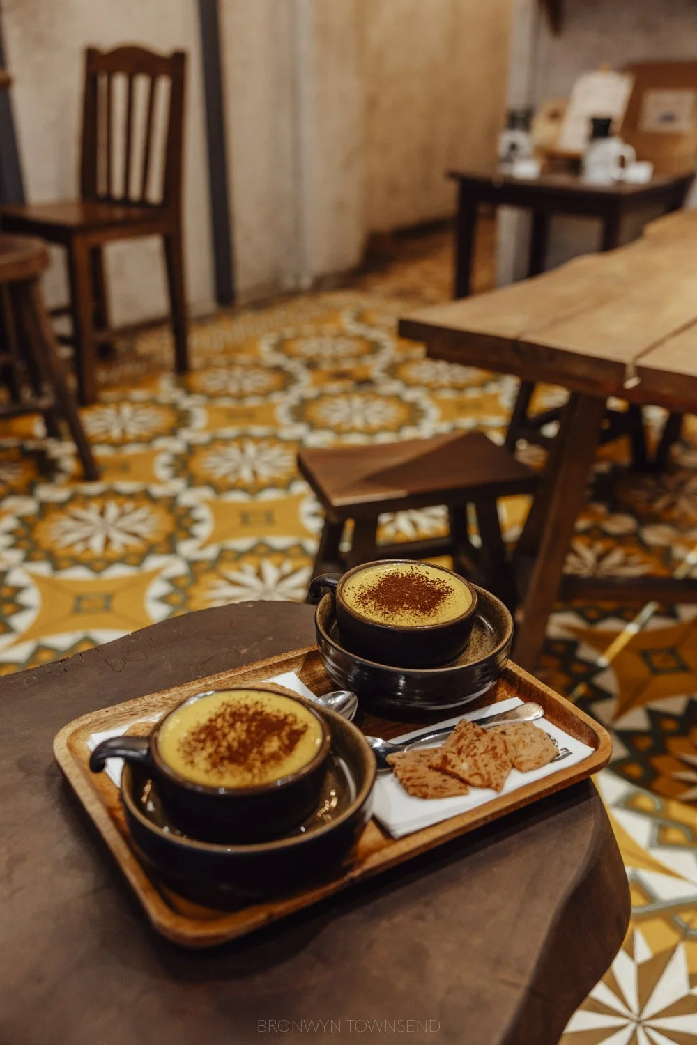 two vietnamese egg coffees on a wooden tray. There are yellow vintage patterned tiles and dark wood furniture in the background