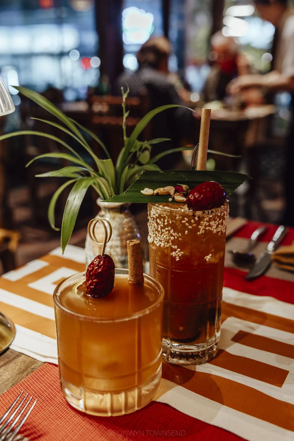 two cocktails in a short and tall glass on a red and white striped table cloth with a plant in the backgroun