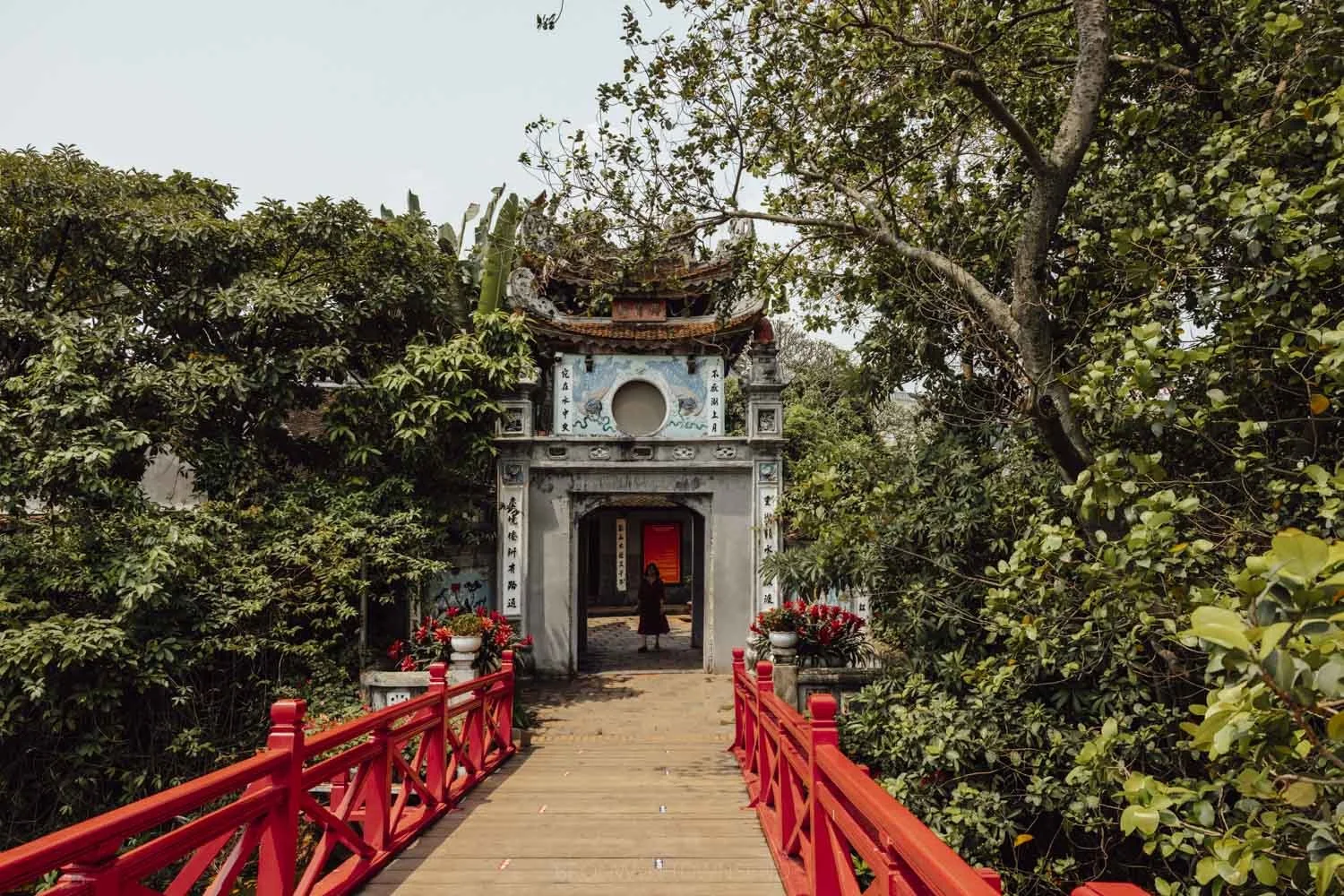 entrance of ngoc son temple from red huc bridge, with temple gate surrounded by large green leafy trees