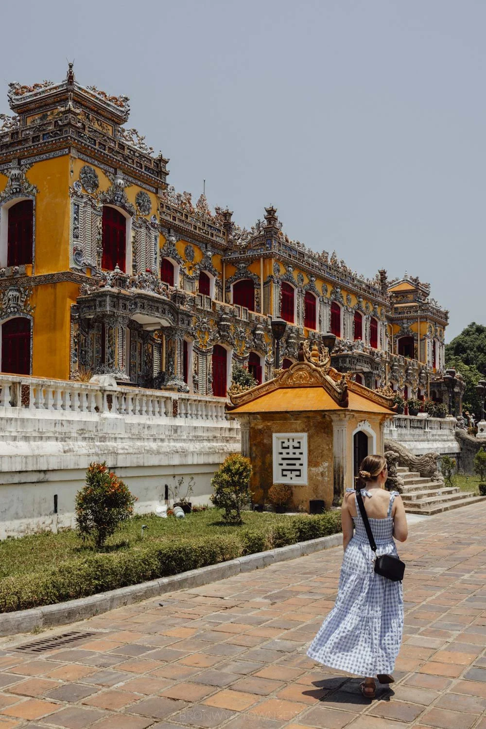 Woman standing with back to camera in a long blue checked dress in front of an orantely detailed yellow and red palace in vietnam