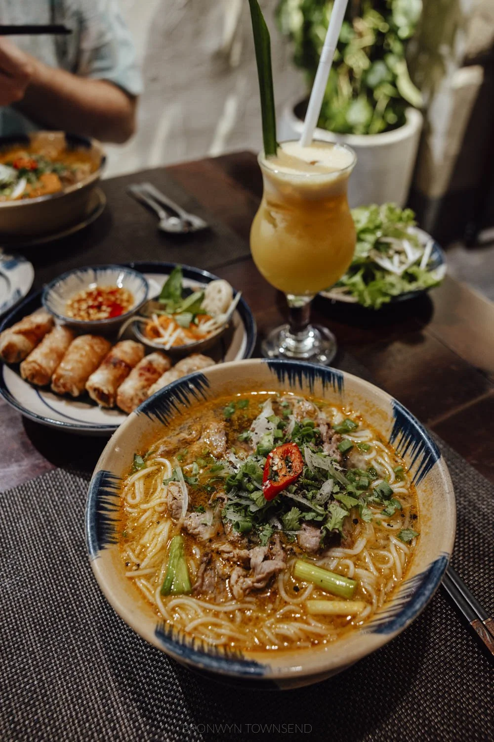 a bowl of bun bo hue, a yellow-shaded noodle and broth dish. There is a mango smoothie and a plate of fried spring rolls out of focus in the background.