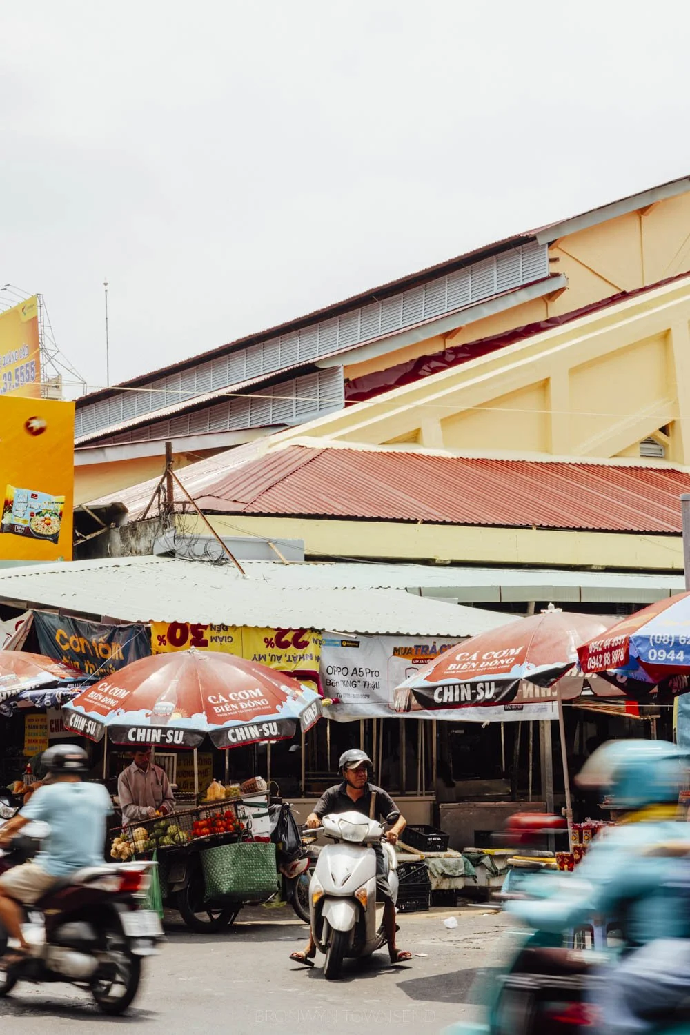 man on scooter outside a yellow and red market building with umbrellas around the exterior