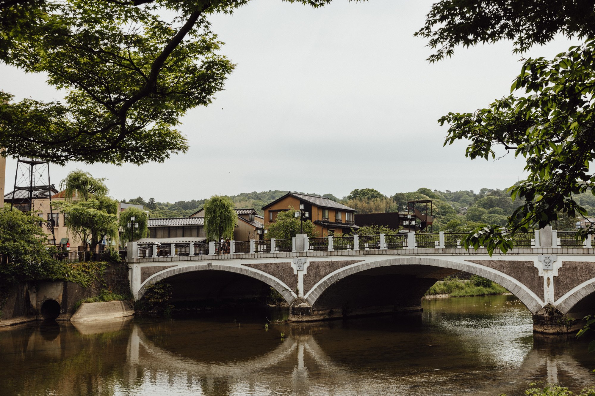 bridge over the asano river with traditional townhouses in the distance, and lush summer foliage of willows and cherry trees hanging over the river banks