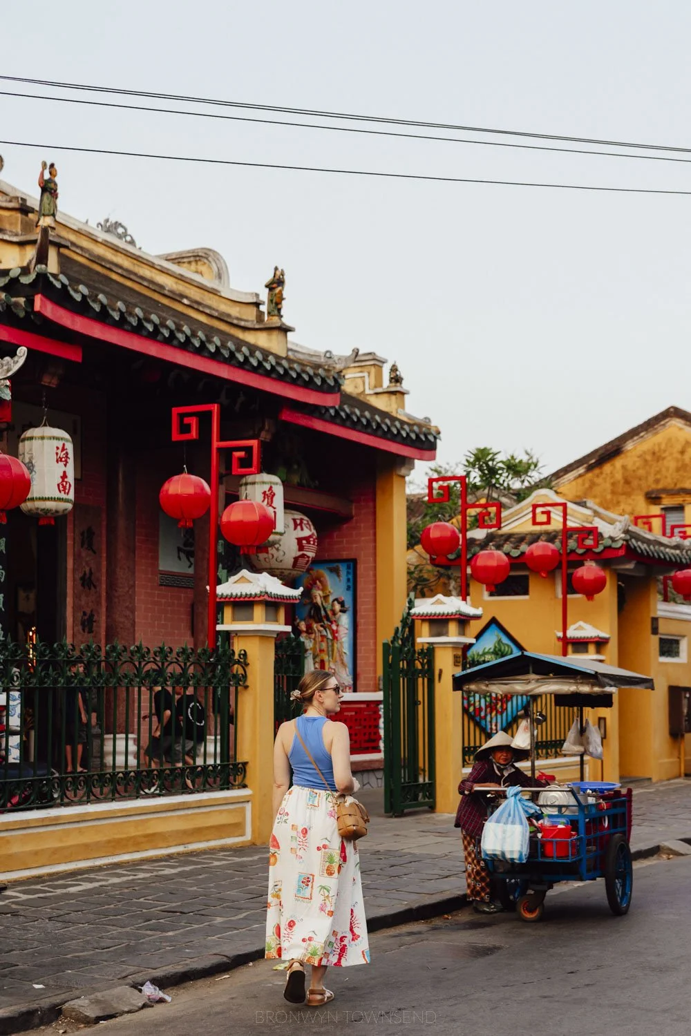 woman wearing a white patterned skirt and blue tank top walking in front of a yellow and red temple building. There is a street vendor selling from a cart in the background.