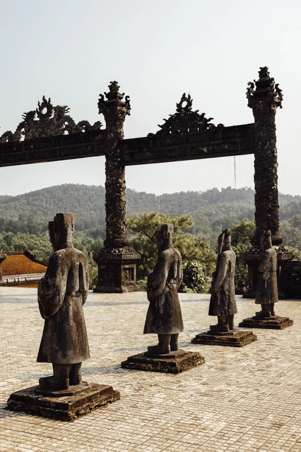 statues made from dark stone and the gates with forested hills in the distance at Khai Dinh's tomb, hue