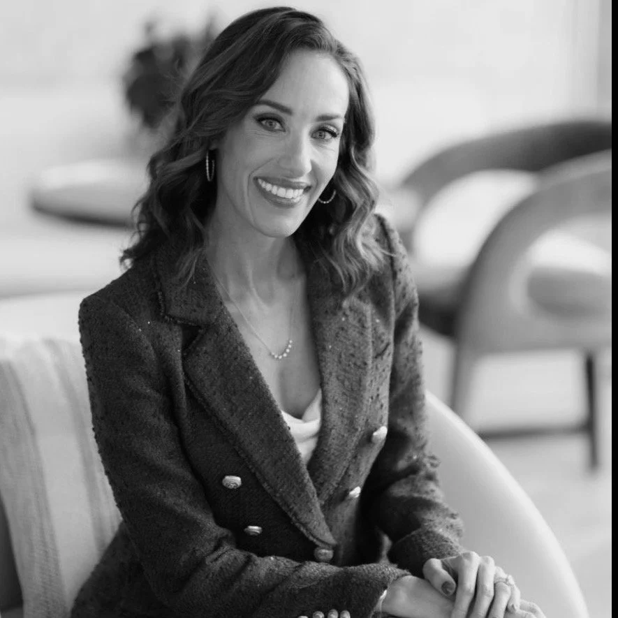 A woman with wavy hair, wearing a blazer and jewelry, smiling while seated indoors.
