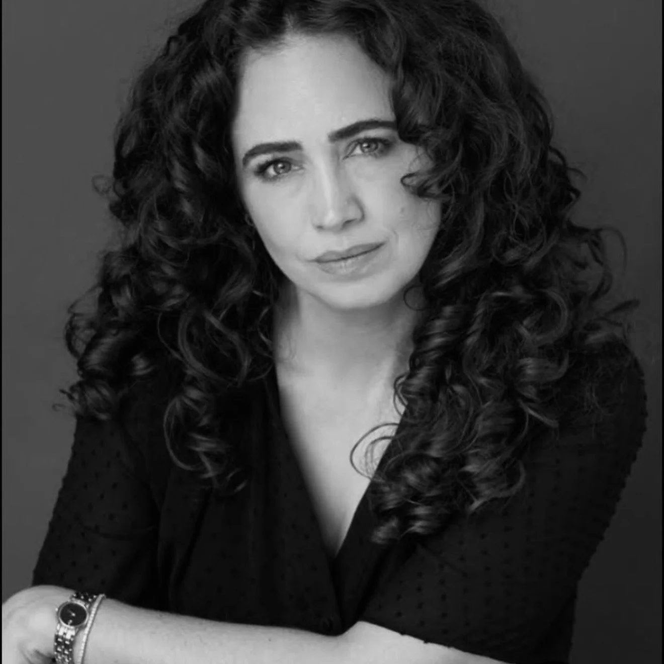 Black and white portrait of a woman with long curly hair, wearing a dark polka-dot top, looking at the camera with a slight smile, with her arms crossed and wearing a watch on her wrist.