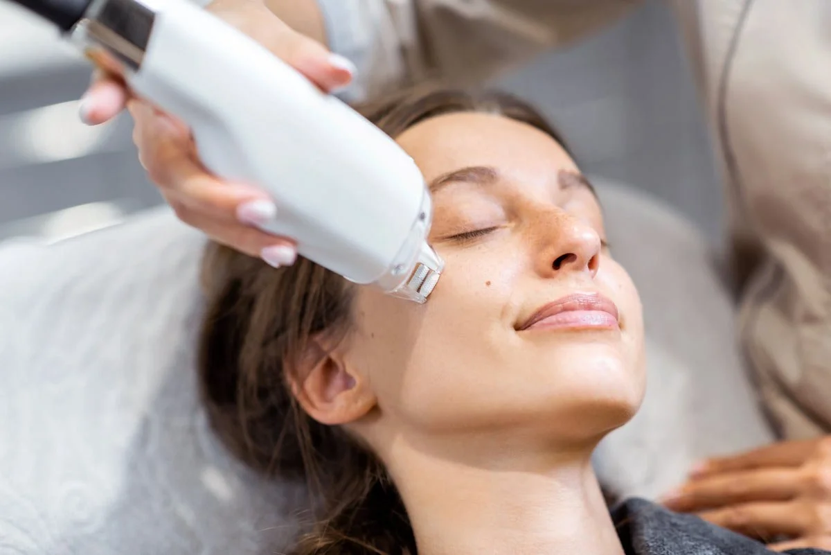A woman receiving a facial treatment, with a device applied to her face, and her eyes closed, appearing relaxed.