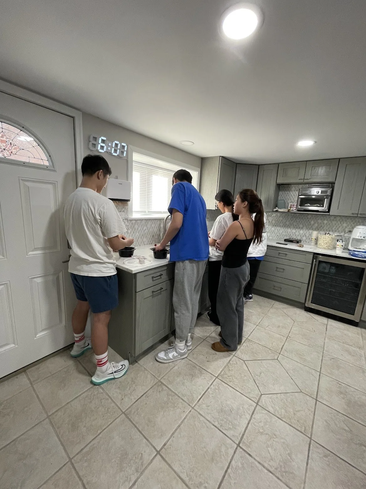 Five people standing at a kitchen counter, preparing food or snacks, with a digital clock displaying 6:07 in the background.