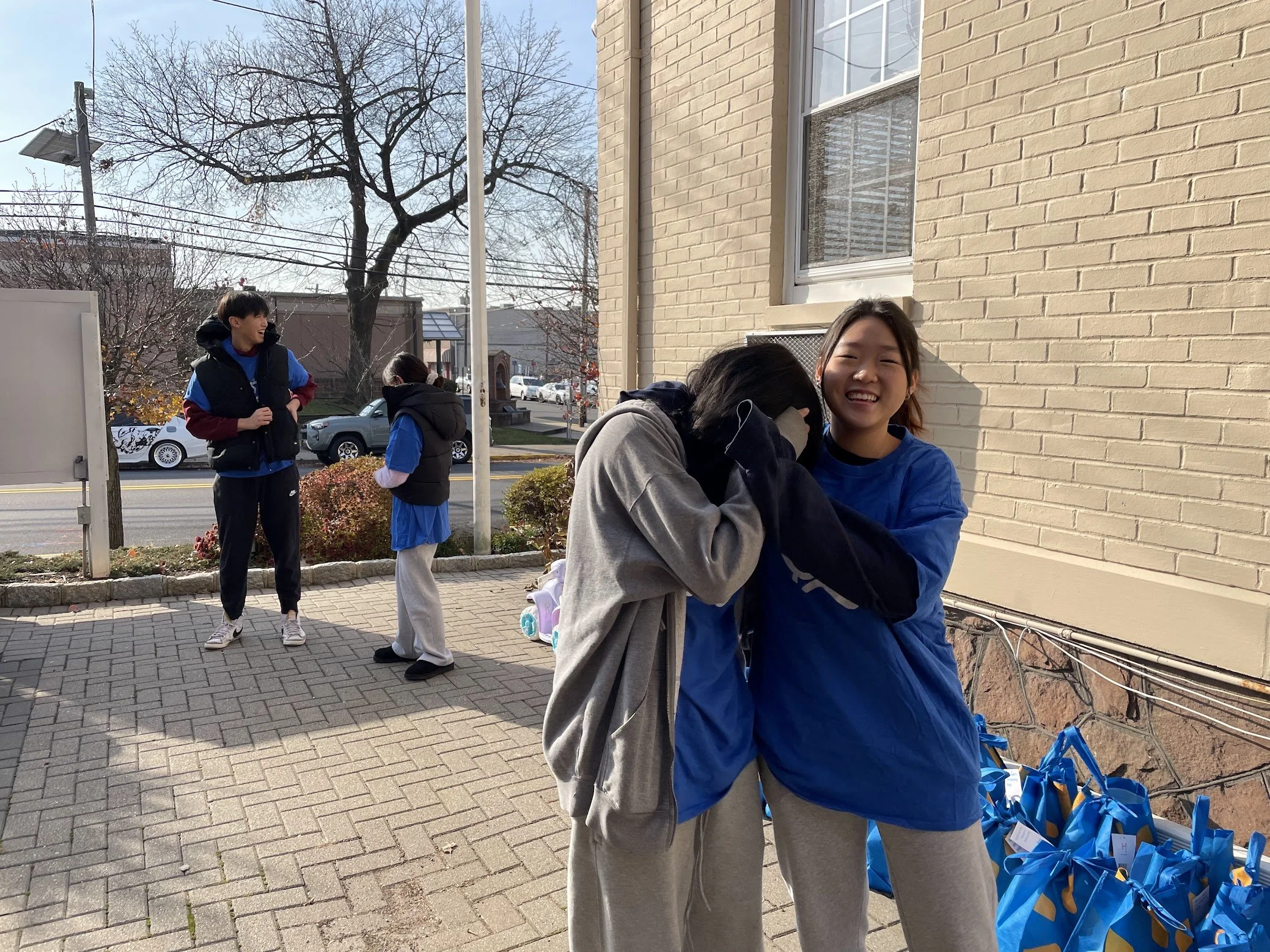 Two young women smiling and hugging each other outdoors near a beige brick building, with several blue gift bags on the ground nearby. In the background, two others are talking near a sidewalk, trees, and parked cars.