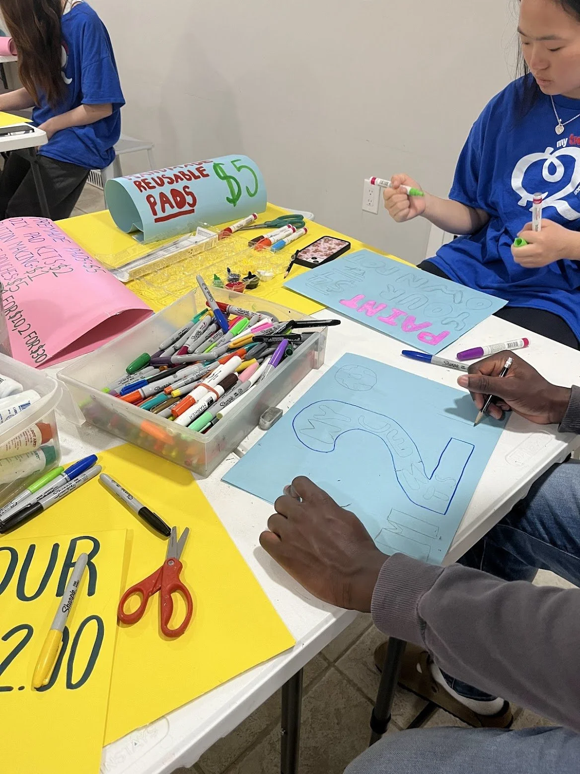 People creating posters and signs at a table for a protest or event, with markers, scissors, and signs reading 'REUSABLE PADS $5' and 'PLANT'.
