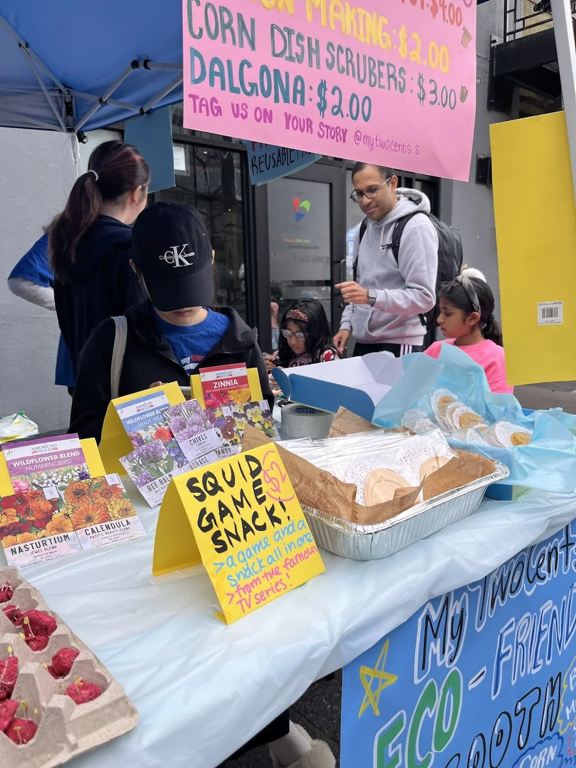 A street market stall selling seeds, popcorn, and a snack labeled 'Squid Game Snack'. Several children and adults are gathered around the stall, which is under a blue canopy. A pink sign lists prices for corn, dish scrubbers, and dalgona, and provides social media information.