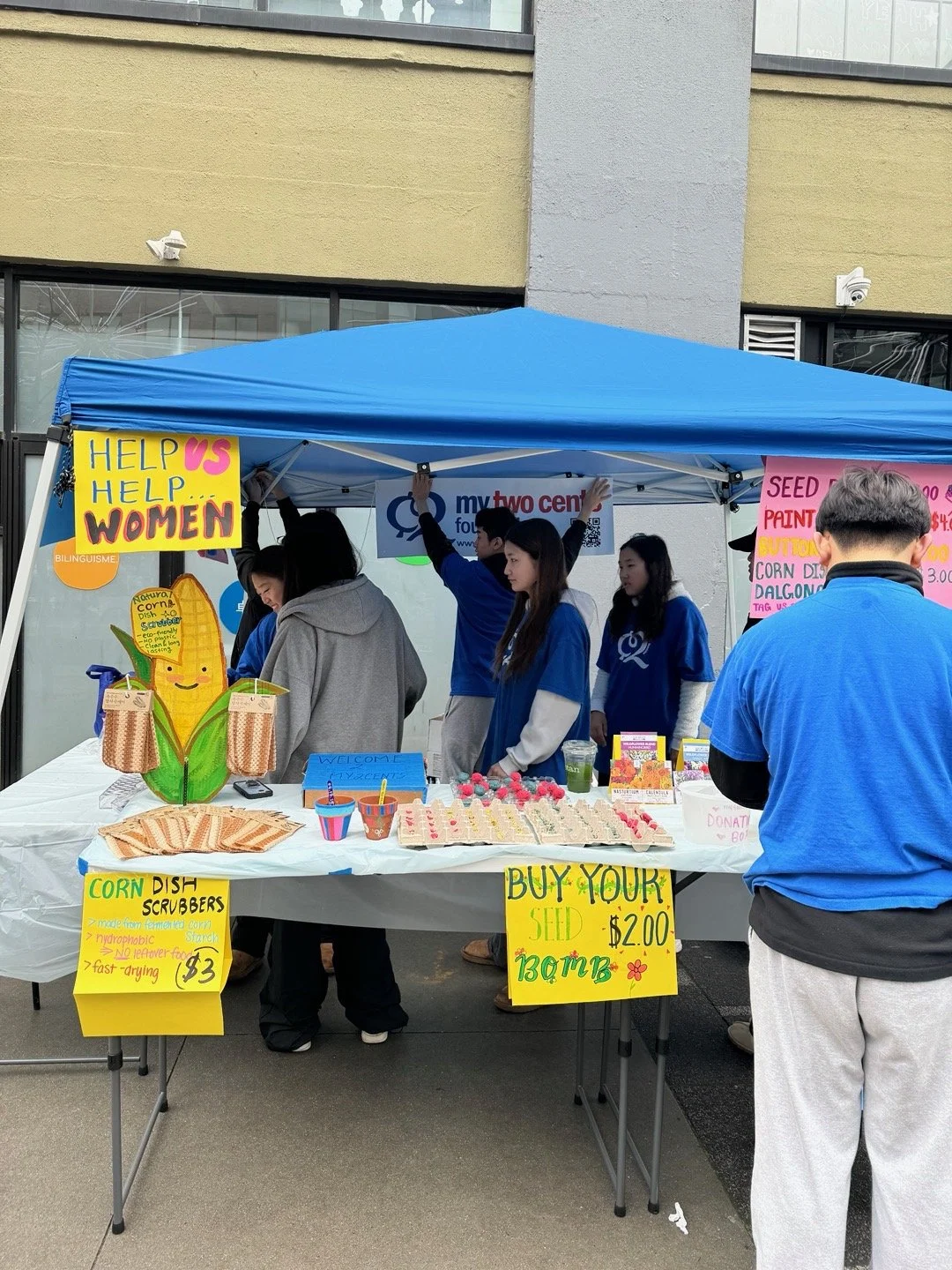 A group of people are at an outdoor booth selling seeds and corn dish scrubbies. The booth has colorful signs, including one that says 'Help Us Help Women' and another encouraging buyers to purchase seed bombs for $2. There are various items on the table and people wearing blue shirts and a gray hoodie. The booth is under a blue tent, and the background shows a building facade with windows.