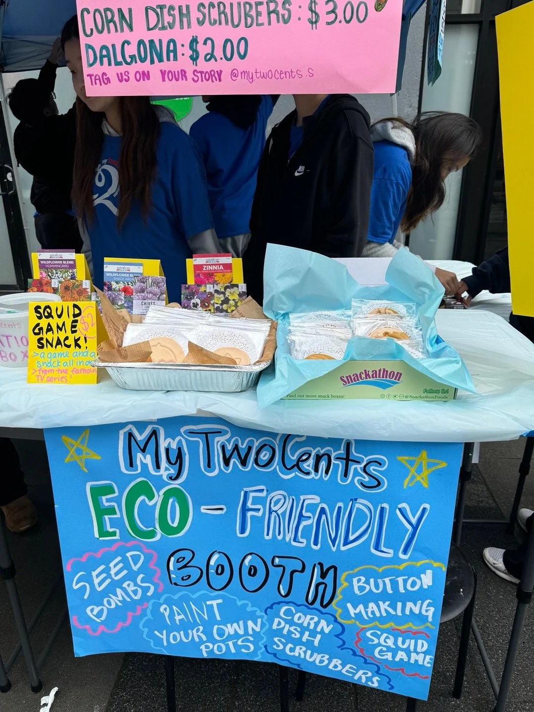 A booth with a colorful poster advertising eco-friendly activities like seed bombs, boat making, and painting your own pots. The booth sells corn dish scrubbers for $3.00 and Dalgona for $2.00. People are gathered behind the table, and the sign includes social media handles and instructions for participation.