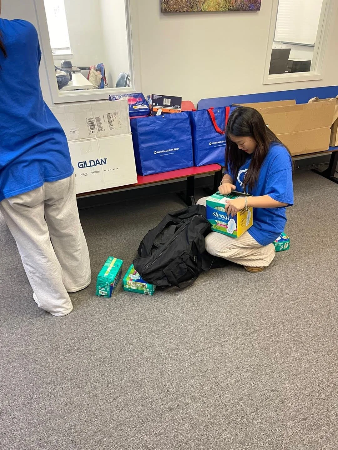 A girl wearing a blue shirt and beige pants is sitting on the floor, opening a package of Always feminine pads. There are boxes of feminine products and a black backpack nearby. A person in a blue shirt and white pants is partially visible, standing nearby. Behind them, there are blue and brown donation bags on a red bench with more packaged products and a window in the background.