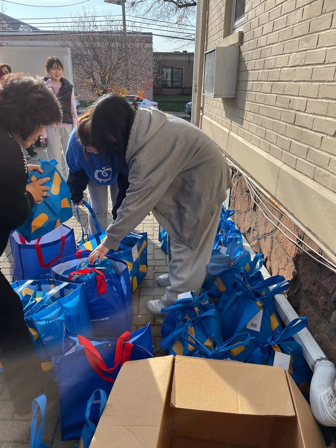 People gather outside to distribute bags, with blue bags with red handles and a cardboard box seen on the sidewalk, next to a building with a brick wall.