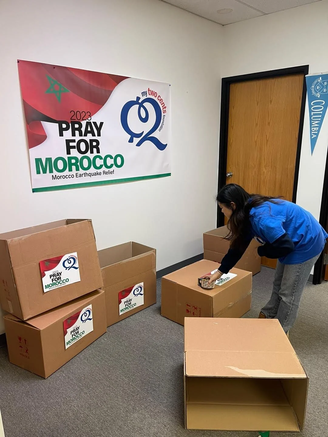 A woman is packing boxes for a humanitarian effort to support Morocco earthquake relief, with a banner on the wall that reads 'Pray for Morocco' and indicates the 2023 Morocco earthquake relief campaign.