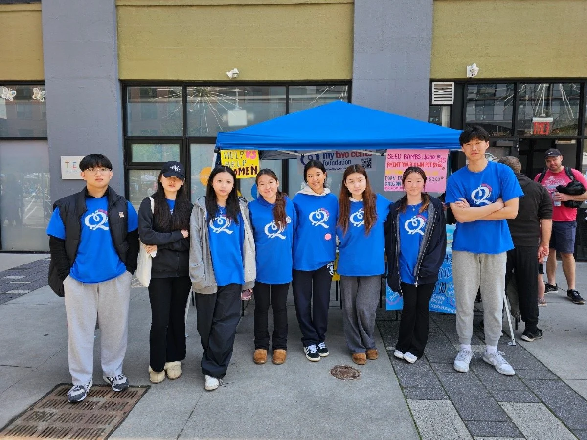 Group of eight young people standing in front of a blue tent for a charity event, wearing matching blue t-shirts with a white logo, with signs on the tent offering help and raising funds.