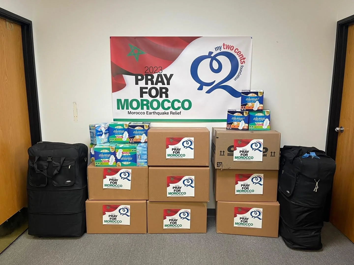Boxes of supplies, including bottled water and toiletries, stacked beneath a banner that reads 'Pray for Morocco' for earthquake relief, with large black and green text and a logo to the right.