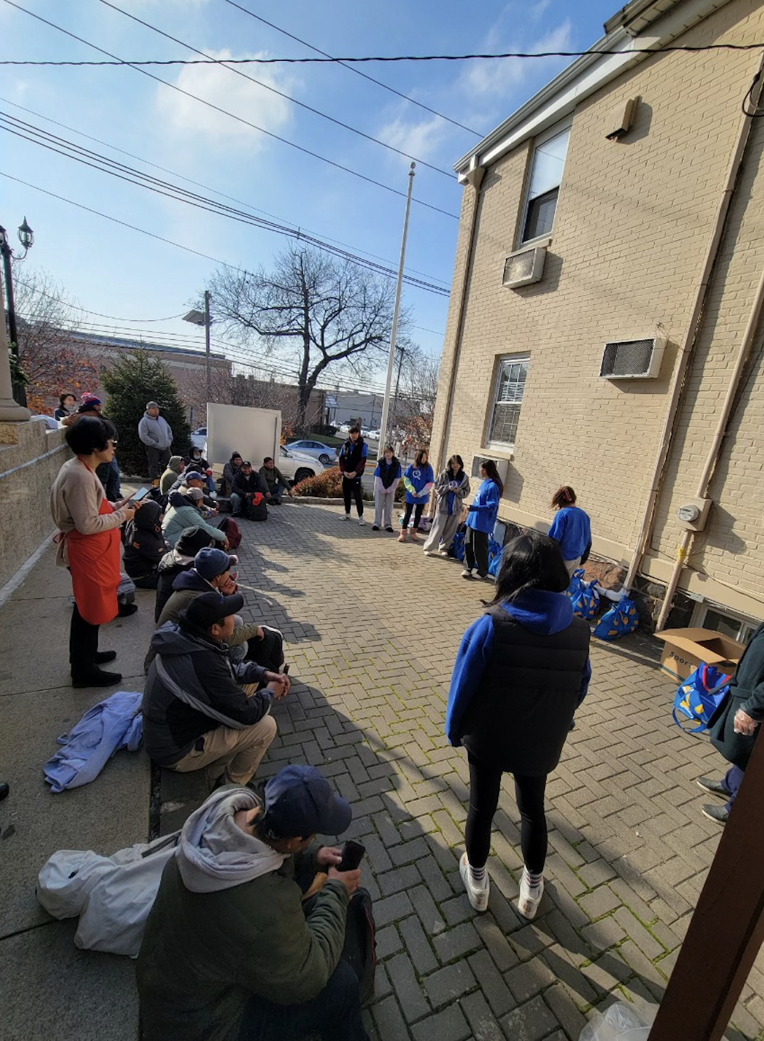 A group of people gathered outdoors in a semi-circle, some seated on the ground and others standing, in front of a beige brick building on a sunny day.