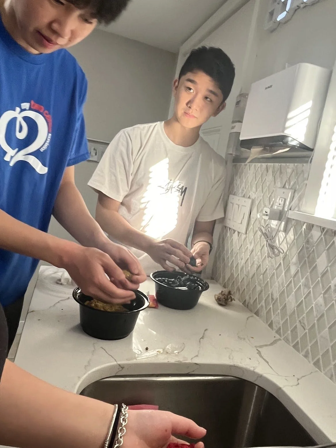 Two young males preparing food in a kitchen. One is wearing a blue T-shirt and the other a white T-shirt, standing by a marble countertop with black bowls.