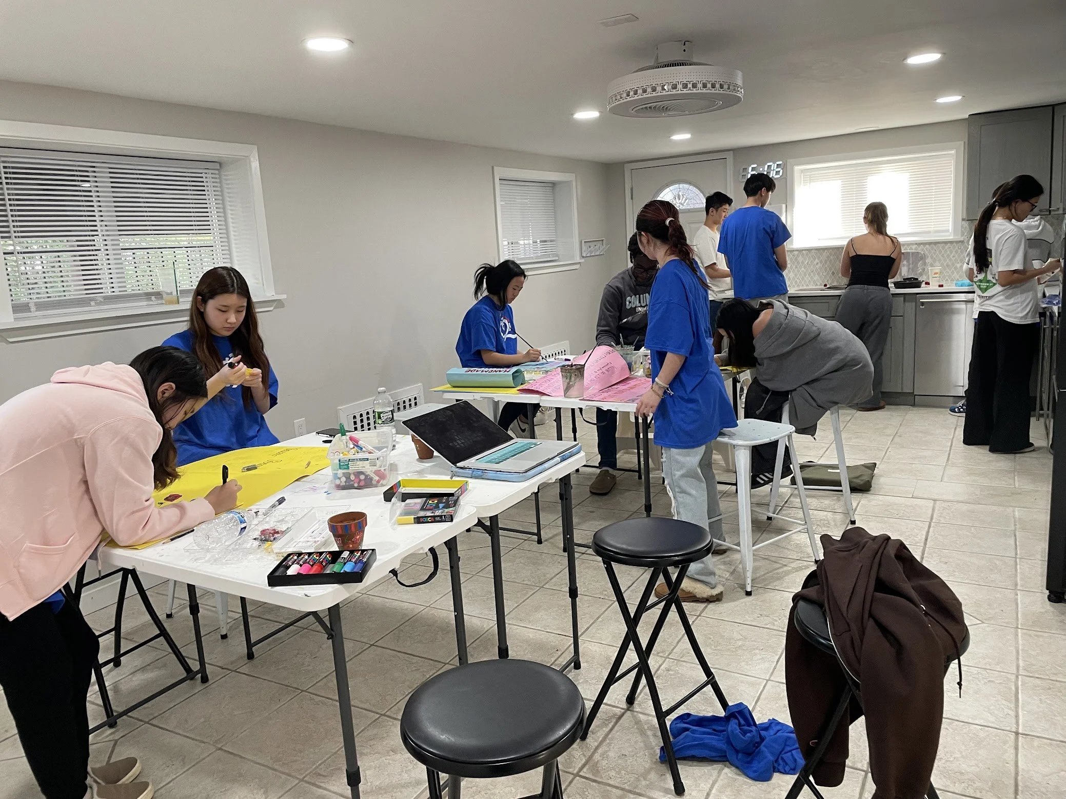 People engaged in arts and crafts in a well-lit room, with tables filled with art supplies, laptops, and notebooks. Some are standing, some are sitting, working on creative projects.