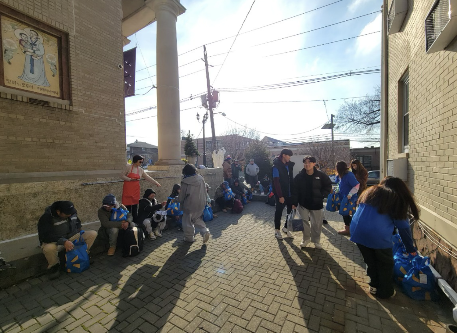 People standing and sitting outside a church, with some wearing face masks, holding blue bags, and talking on the phone, in a sunny day.